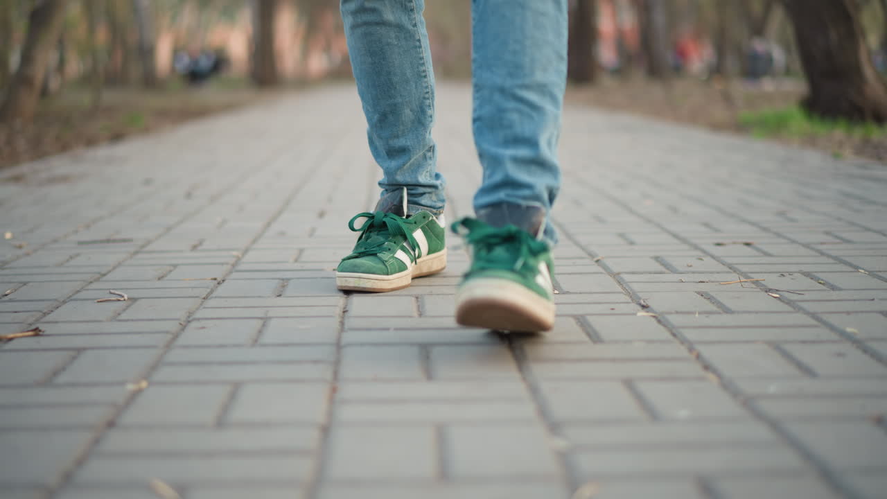 Primer plano dinámico de zapatillas verdes moviéndose sobre un camino embaldosado de la ciudad, rápidos toques y arrastres de los dedos de los pies, pantalones vaqueros, animado parque de fondo, joven bailarín callejero practicando pasos, energía de fin de semana, enfoque superficial