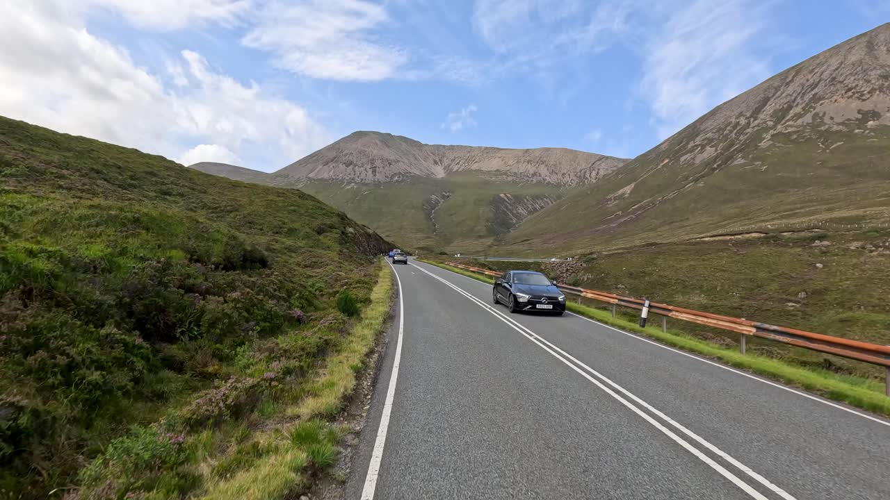 A black car travels along a winding mountain road in daylight, surrounded by green hills and dramatic skies, captured in a smooth forward-moving shot