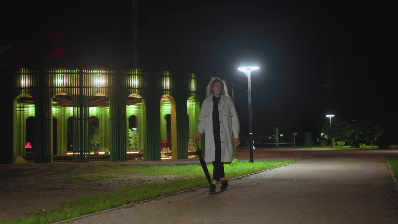 Woman dressed in white coat carrying umbrella walking calmly through urban park at night, illuminated by bright streetlights, green pavilion glowing in background