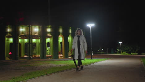 woman dressed in white coat carrying umbrella walking calmly through urban park at night, illuminated by bright streetlights, green pavilion glowing in background
