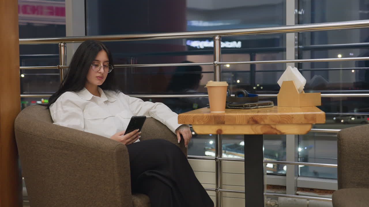 Curious woman with glasses seated comfortably in indoor lounge holding phone while gazing with interest, coffee cup, tablet, and headphones rest on wooden table in front near window with city lights