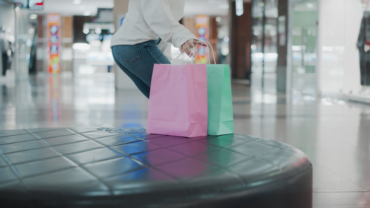 쇼핑몰에서 가죽 좌석에 앉아있는 여성은 가게와 밝은 조명을 특징으로하는 흐릿한 소매 배경으로 떠나기 전에 민트와 분홍색 쇼핑 가방을 집어 들고 있습니다.