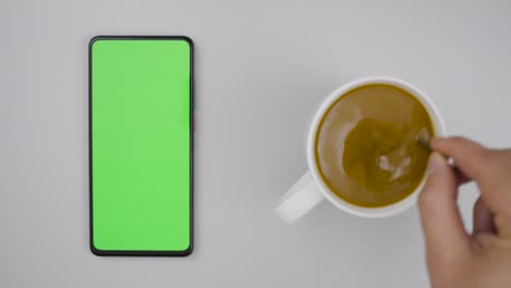 smartphone with green mock-up screen business concept. person hand stirring coffee with spoon on table. slow motion. man hand cup of hot coffee and using smartphone watching green screen top view.