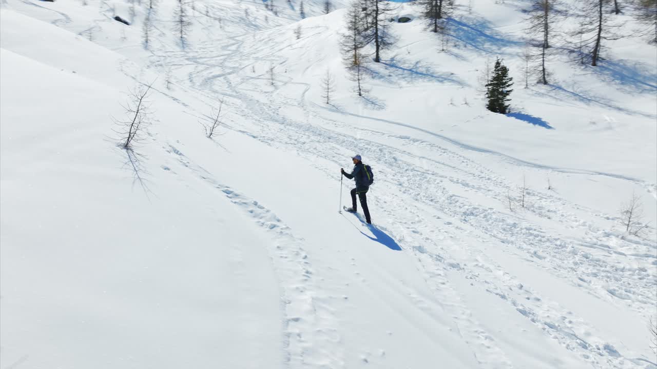 Person snowshoeing on pristine snow, leaving footprints, sunny winter day, long shadow, Cheneil, Italy. Aerial drone orbiting at low altitude