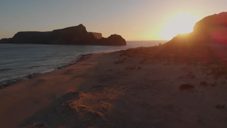 Toma-Aérea-Hacia-Atrás,-Luces-De-Puesta-De-Sol-En-La-Playa-De-Calheta,-Islote-Como-Fondo