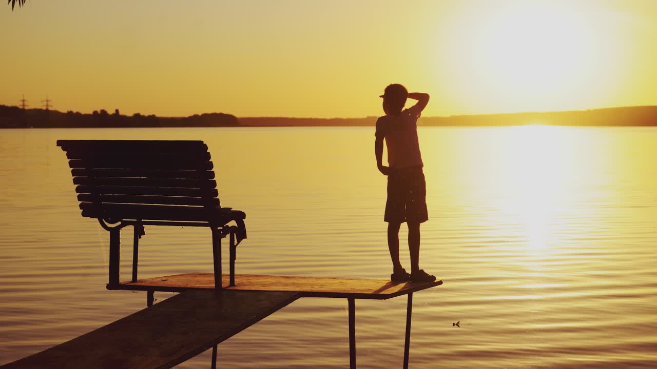 A little kid is standing on the masonry near the bench. The guy is looking around the area at sunset near the lake in the summer.