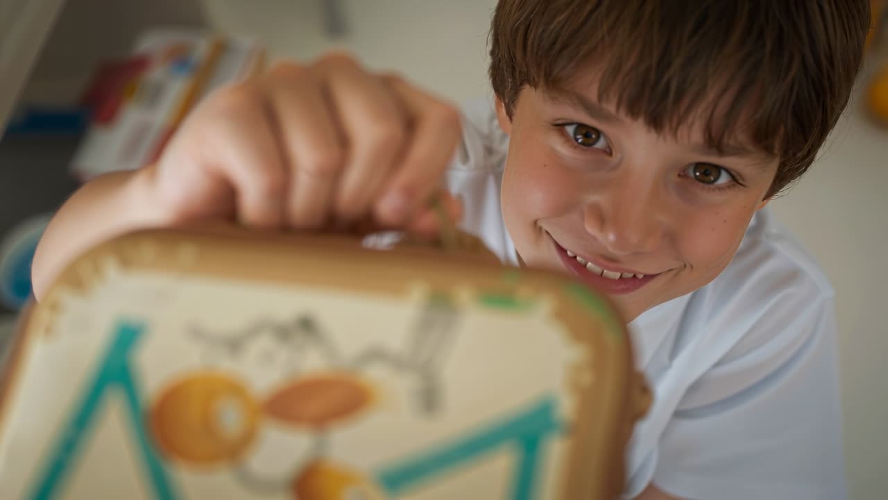 Niño con camiseta blanca levantando una lonchera de dibujos animados en casa, ofreciéndosela a un padre, con una mano anillada ayudándole.