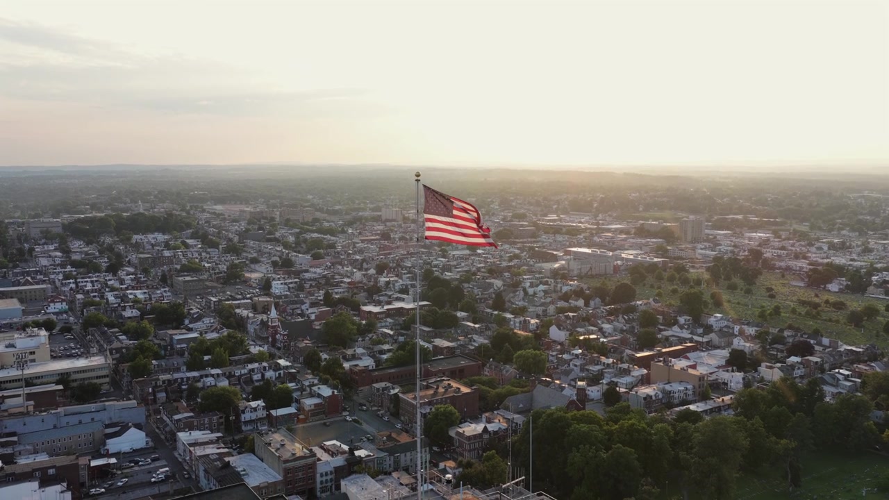 Enorme bandiera americana sventola in cima a un alto pennone bianco sopra i tetti del centro di Allentown, fitte case a schiera e il campanile di una chiesa che si estendono su uno skyline sfocato al tramonto in Pennsylvania. Primo piano aereo con drone