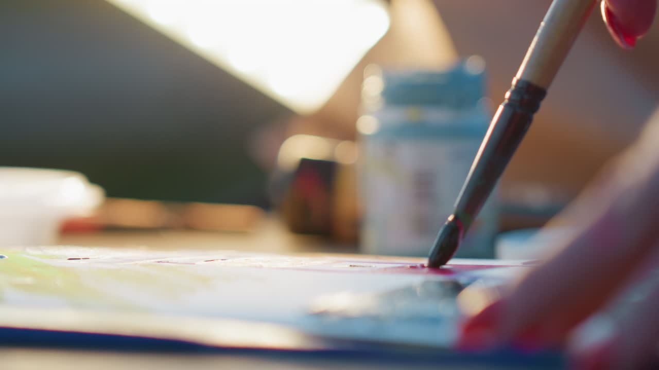 close up artist hand dipping brush into paint and applying colorful strokes on paper with blurred background bokeh lights capturing creative motion and texture in soft glowing evening light outdoors