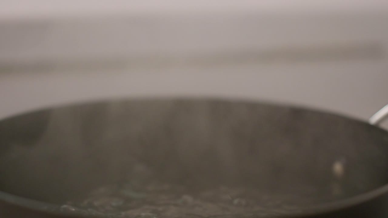 Simple, extreme close up shot of man's hand lifting the lid of a pot of boiling water sitting on a gas cooktop, and letting steam escape