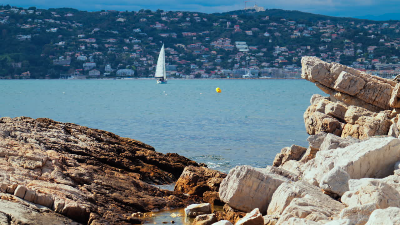 Rocks on the shore wit a sail boat moving on the sea in Juan-les-Pins, France with the city and the mountain on the background