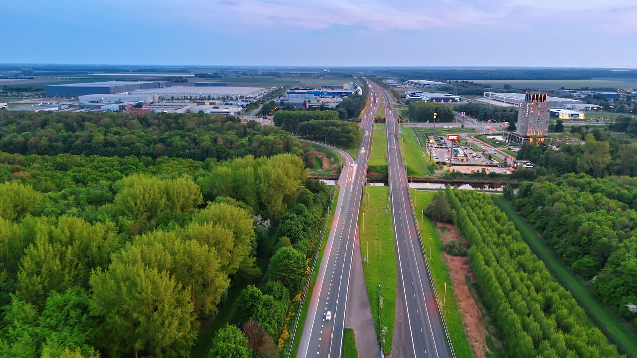 Winding road through lush landscape. Aerial view captures a highway winding between vibrant green forests and open fields near urban development at dusk