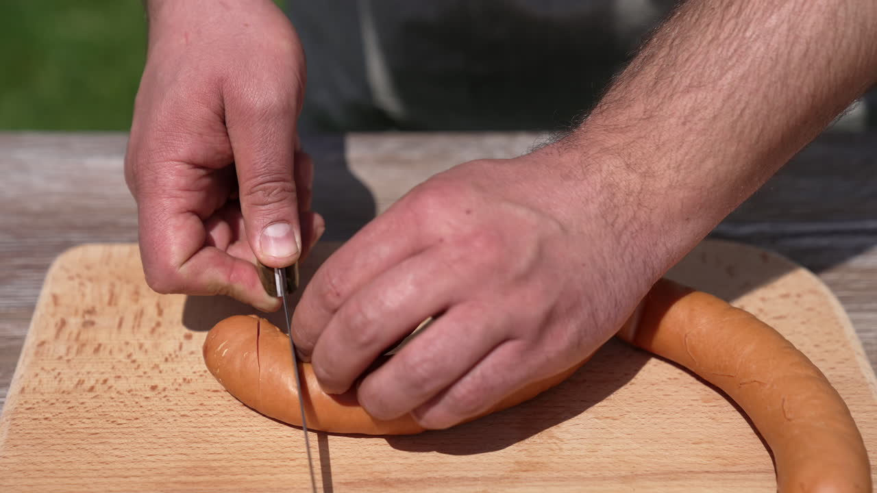 Cooking sausages in the open air. Man making an incision on sausages with a sharp knife before frying on a grill. Close-up.