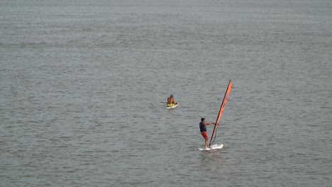 Chico-Haciendo-Windsurf-En-El-Río-Han-Con-Un-Hombre-Sentado-En-Una-Tabla-De-Surf-En-El-Fondo