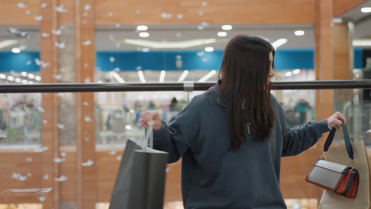Female shopper in casual outfit stands near glass railing of modern mall, gazing at colorful retail store below while waiting for friend after shopping, surrounded by soft lights and flying decor
