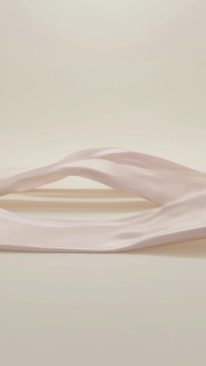 Video verticale: Centro che si solleva, facendo ondeggiare un nastro rosa pallido su un set beige, con luce soffusa.