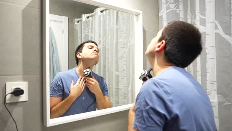 Man-shaving-off-beard-with-electric-shaver-plug-to-socket,-mirror-reflection