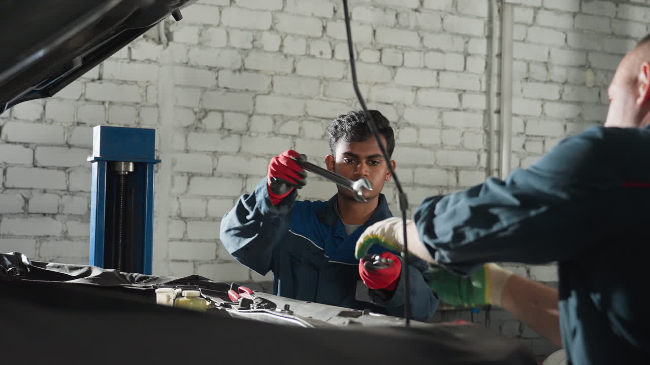 primer plano de dos ingenieros en uniformes azules trabajando en el motor del coche, intercambiando herramientas y haciendo ajustes en un taller profesional de automóviles con iluminación industrial