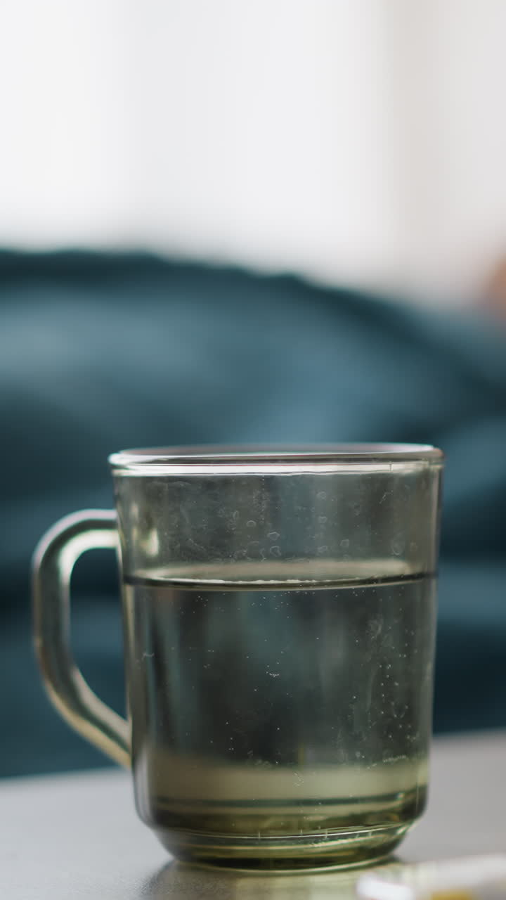 Water inside glass cup on table with thermometer and blurred background showing restless person on couch, dim light indicating a sick day, recovery, and care at home