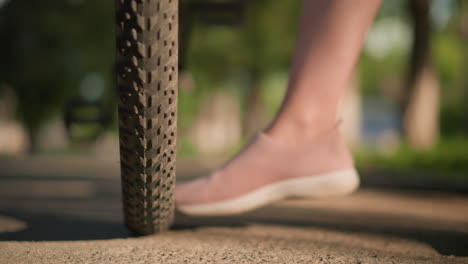 close-up of someone's legs wearing pink sneakers kicking a bicycle tire to check air pressure, the scene captures lush greenery and trees in the blurred background