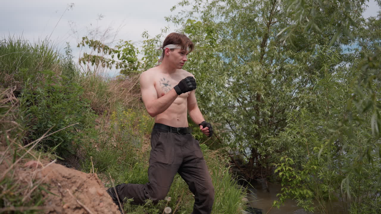 Young soldier standing shirtless near riverbank with clenched fists and strong leg stance, rehearsing focused punches under cloudy sky in lush outdoor environment surrounded by grass and trees