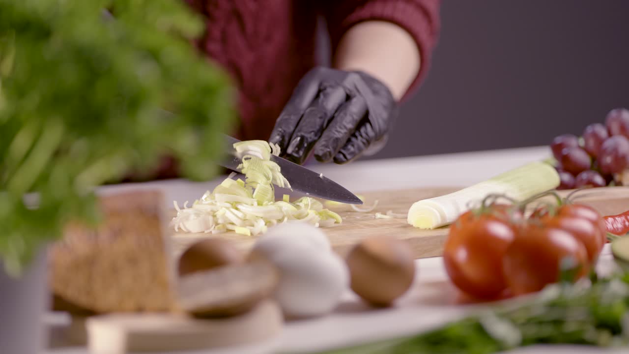 A chef expertly slices a fresh leek into thin rings on a wooden board in slow motion. The precision of the sharp knife is highlighted, while black gloves maintain cleanliness. Fresh ingredients