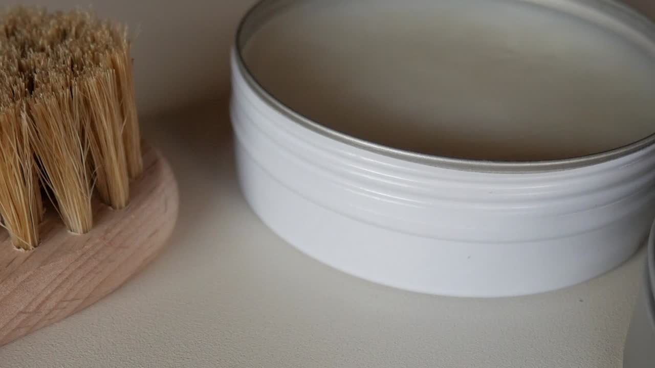 Close-up of a wooden shoe polishing brush and a white wax container placed on a light surface under natural daylight