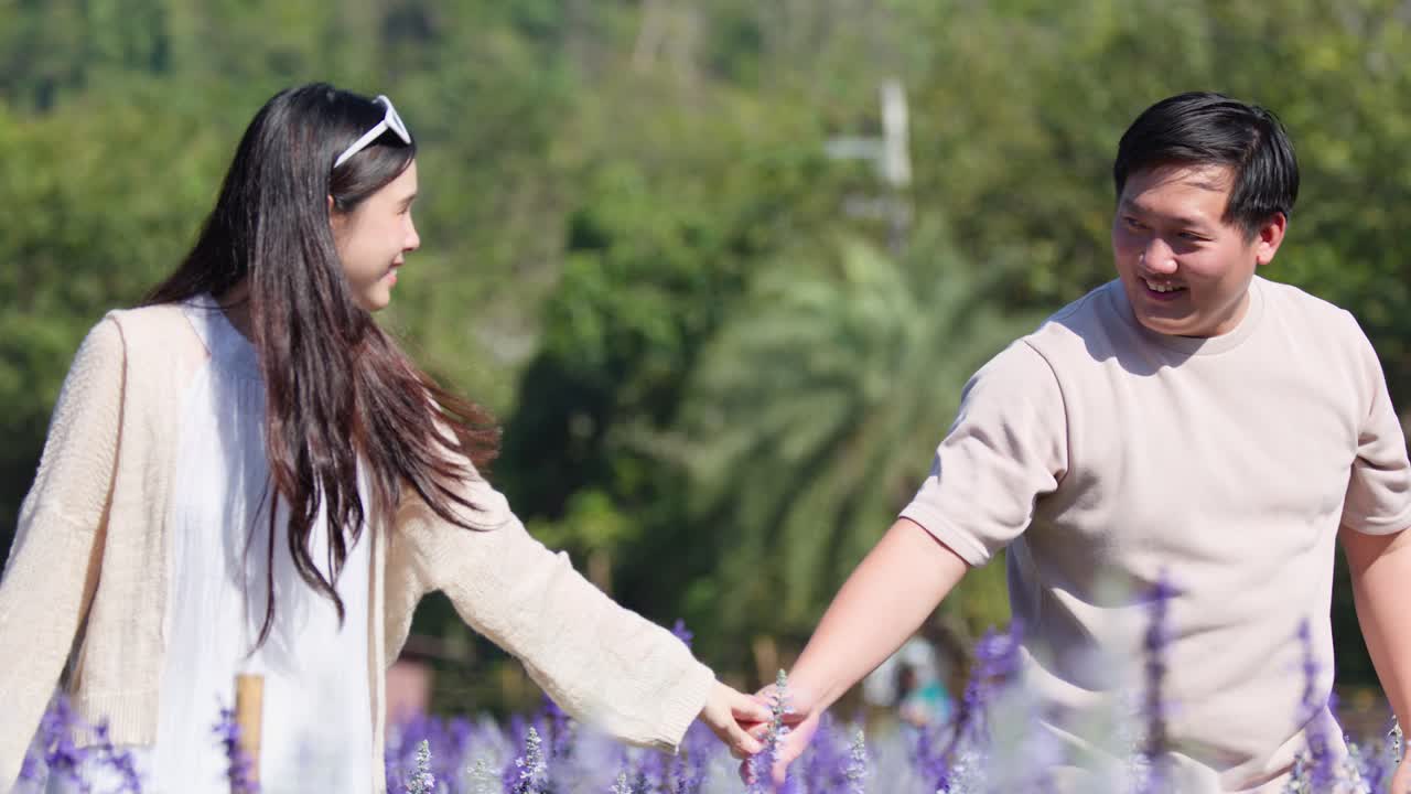 Smiling couple walks hand in hand through sunny purple flower garden, medium tracking shot