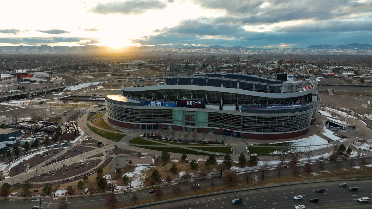 브롱코스 스타디움 이전의 마일 하이 스타디움에서 상징적인 임파워 필드에서 멀리 날아가는 드론과 일몰 동안 메인 스트리트 고속도로와 시내 덴버 시티 뷰 배경을 통해 운전하는 자동차를 드러내는 것