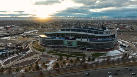 Drone-Volando-Lejos-Del-Icónico-Campo-De-Empoderamiento-En-El-Estadio-De-Una-Milla-De-Altura,-Anteriormente-Estadio-De-Los-Broncos,-Y-Revelando-Autos-Conduciendo-A-Través-De-La-Carretera-Principal-Y-El-Fondo-De-La-Vista-De-La-Ciudad-De-Denver-Durante-La-Puesta-De-Sol