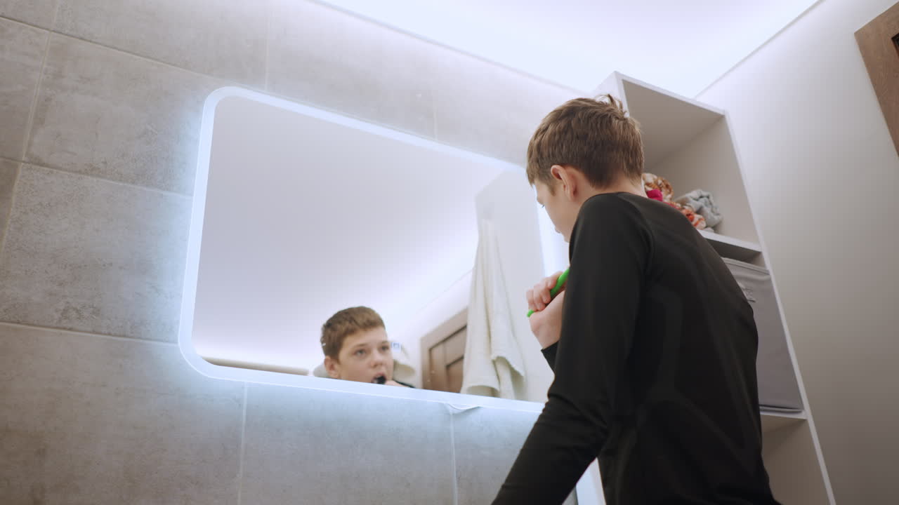 Ragazzo bianco che controlla il riflesso in uno specchio da bagno a LED mentre si china sul lavandino, con lo spazzolino in mano, in un ambiente di piastrelle in penombra, atmosfera notturna introspettiva, con moderno mobile lavabo e articoli da toeletta visibili.