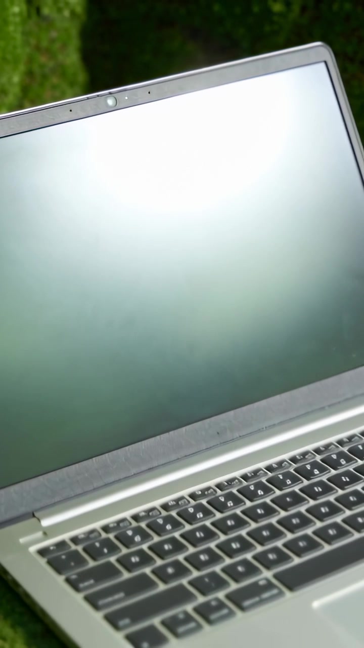 Vertikal video: Nærbillede starter med kameratilbagetrækning, der afslører en sølvfarvet bærbar computer på græs, der viser et webcam