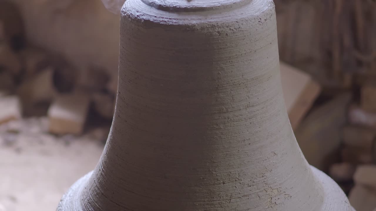 Camera pans down across surface of freshly cast bell mold still emitting smoke and heat, showing fine clay textures and the recognizable flared profile of traditional foundry production, slow motion