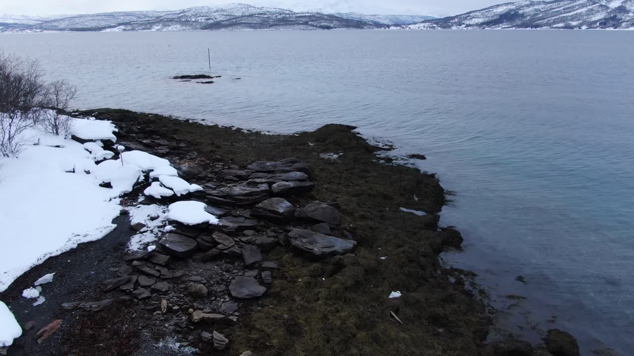 дрон в тромсо, летящий к морю из заснеженной местности с горами, полными снега зимой в норвегии