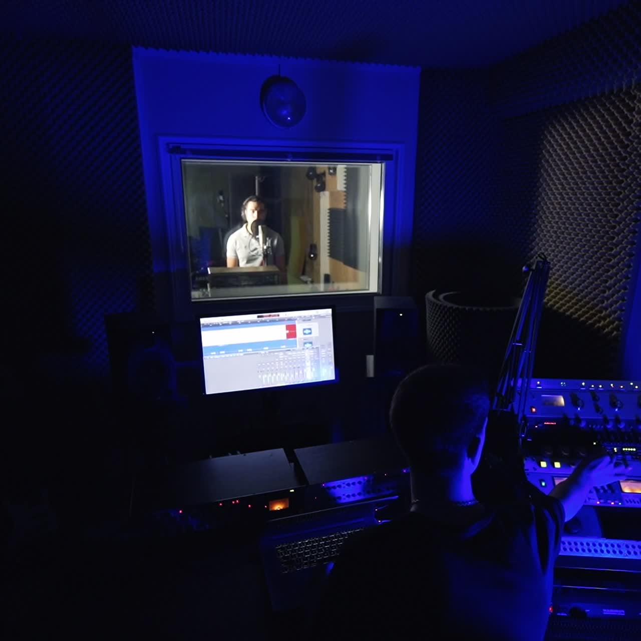 Music engineer sitting in dark studio in front of sound board. Man singing in the next room behind the glass. Top view