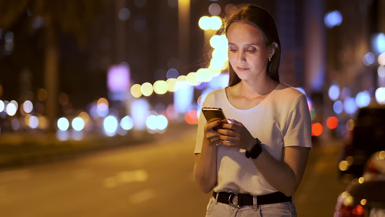 A beautiful young girl in the summer in a big night city keeps a smart phone in shell and writes a message reads e-mail and communicates in chat rooms and social networks keeps a blog looks at the phone screen