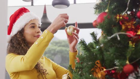 video of happy biracial woman in santa hat smiling and decorating christmas tree at home