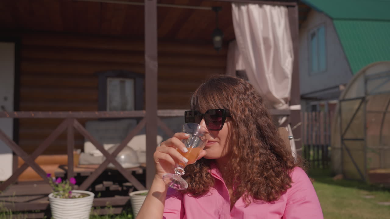 Orange Beverage Savored In Peaceful Garden Setting, Serene Backyard Scene With Lady Drinking Juice And Gentle Wind, Casual Summer Moment Featuring Woman Relaxing On Porch With Refreshing Citrus Drink