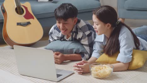 asian teen couple using a laptop while lying on carpet on the floor at home. watching movie with snacks, having video call, speaking, and enjoying time together