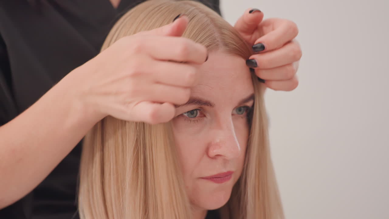 Cliente bionda che riceve un trattamento lucidante per capelli, primo piano di una stilista che applica il gloss con precisione sui capelli biondi, vista dettagliata di una professionista che applica il gloss sui capelli biondi sotto una protezione rosa