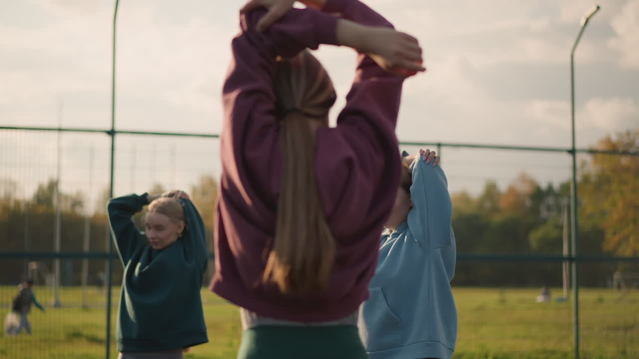 молодые дамы занимаются фитнес-упражнениями, делают растяжки рук на открытом воздухе с барным забором и зеленью на заднем плане человек, идущий за пределами двора