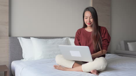 A-young-Asian-woman-sits-cross-legged-on-a-hotel-bed,-with-a-laptop-in-her-lap