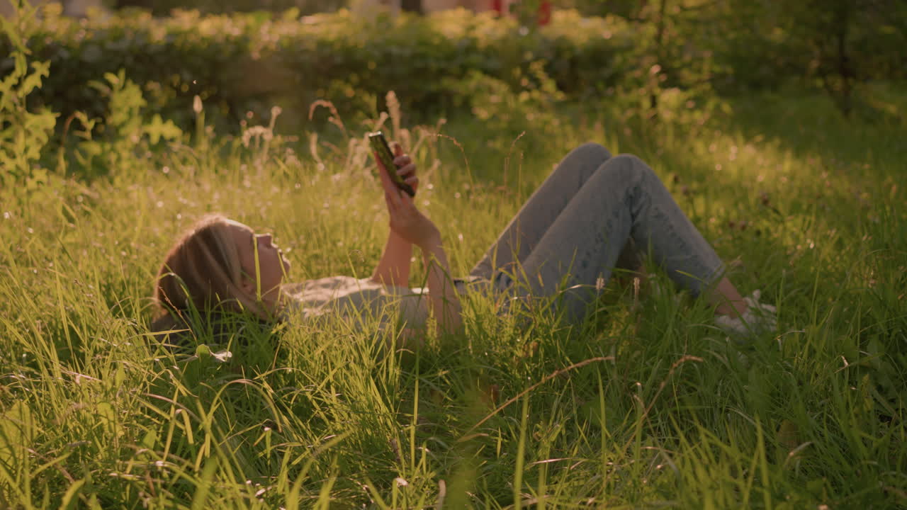 женщина лежит на травяном поле в расслабленном положении, используя смартфон под теплым солнечным светом, в окружении высокой зеленой травы и естественной зелени