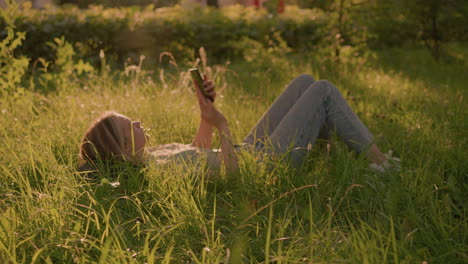 mujer acostada en un campo de hierba en una posición relajada, usando un teléfono inteligente bajo la cálida luz del sol, rodeada de alta hierba verde y vegetación natural