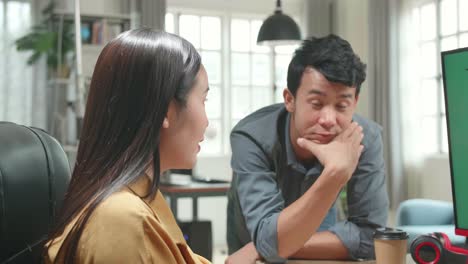 asian female works with her male colleague on a project on her personal computer with mock-up green screen. they work in a creative loft office