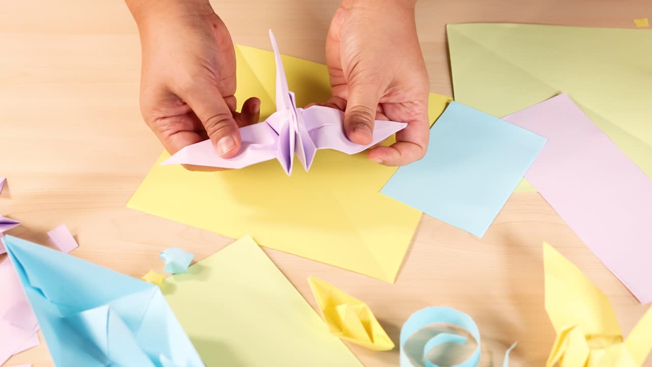 Person skillfully folds pastel origami crane, surrounded by colorful paper crafts, under soft lighting
