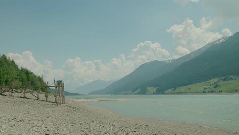 Full-shot,-Wooden-Bridge-on-the-shore-of-Reschensee-in-Italy,-the-Melcesine-mountain-range-in-the-background