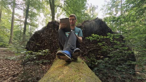 A-man-is-swiping-up-and-down-at-his-tablet-computer-while-he-is-sitting-at-a-tree-trunk-at-a-sunny,-beautiful-day-in-a-green-forest