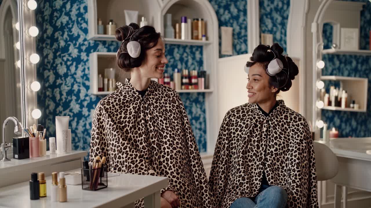 Two cheerful young women with hair curlers wearing leopard print capes are sitting in front of a mirror in a vintage beauty salon, chatting and smiling while waiting for their hair treatment