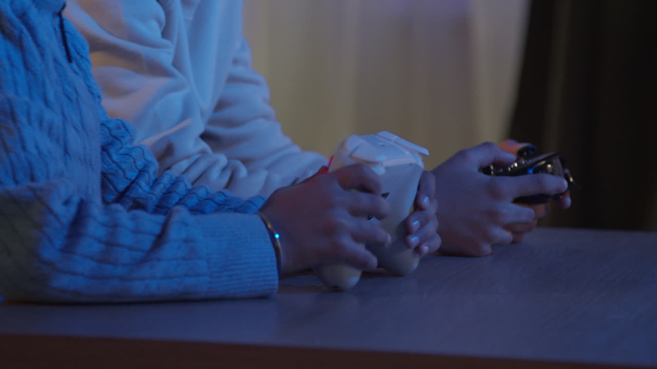 Close Up On Hands Of Two Young Boys At Home Playing With Computer Games Console On TV Holding Controllers Late At Night 2